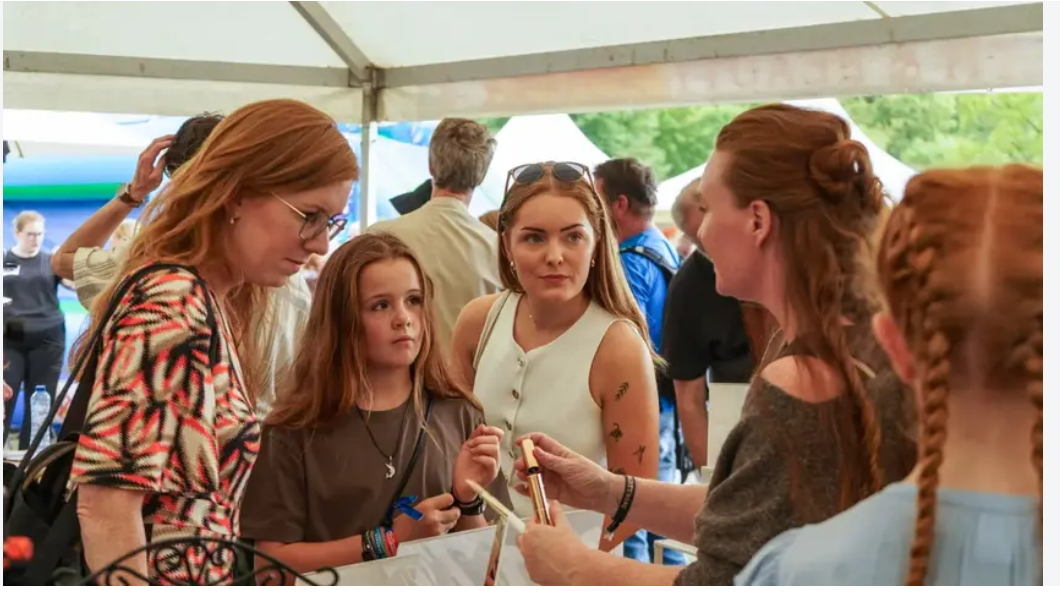 A group of young women with red hair interact at a booth under a white tent at an outdoor event. Eva, founder of Hey Redhead, a mascara brand for natural beauty is at the center of the frame